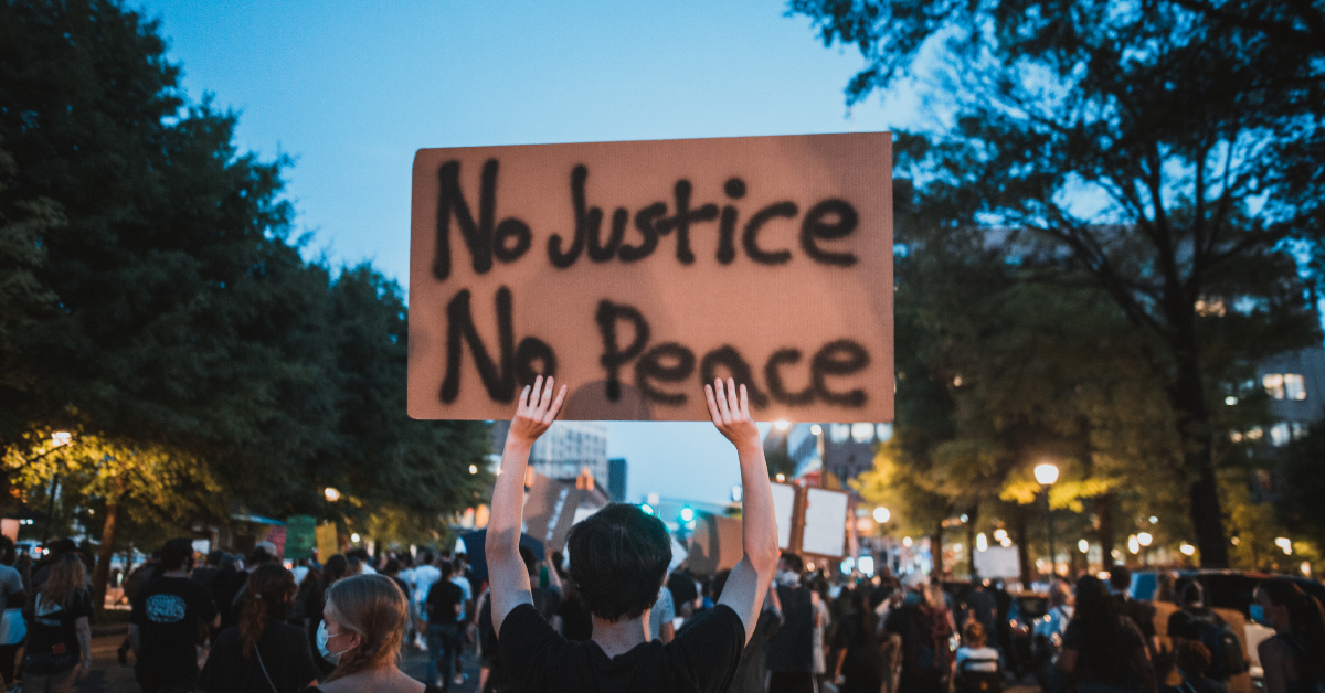 Sign being held over a crowd at an activism rally, which reads No Justice No Peace.