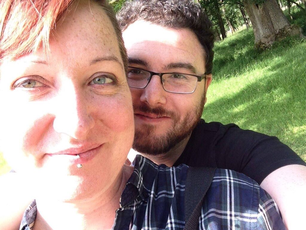 A smiling close-up of Lora O'Brien and Jon O'Sullivan, founders of the Irish Pagan School, enjoying a sunny day in Phoenix Park, Dublin. Lora, with short red hair and a lip piercing, stands in front, while Jon, wearing glasses and a beard, smiles from behind. The background shows green grass and trees, creating a peaceful, natural setting.